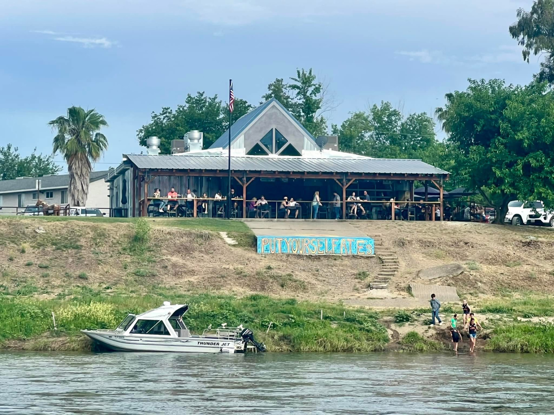 E's Locker Room seen from across the Sacramento River, with guests seated under the covered patio and a 'Put Yourself at E's' mural on the riverbank.