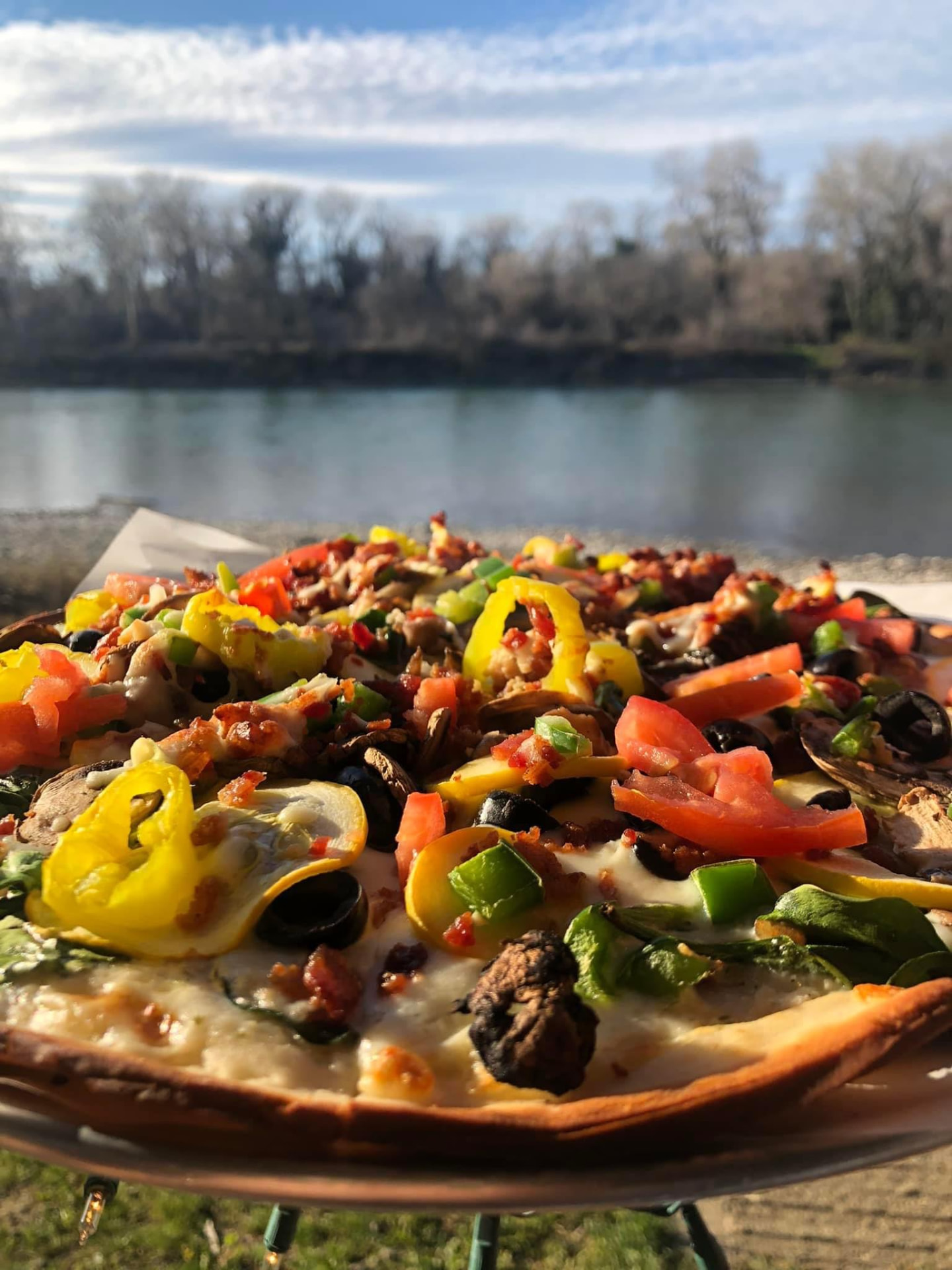 A loaded veggie pizza with peppers, olives, mushrooms and tomatoes, with the river behind it.